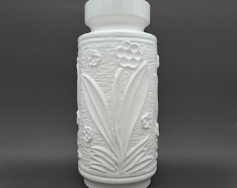 May include: A white ceramic vase with a textured floral design. The vase has a cylindrical shape with a wide base and a flared top. The embossed pattern includes stylized flowers and leaves, adding a decorative element to the piece.
