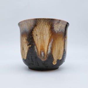 May include: A ceramic cup featuring a dark brown base that transitions into a lighter tan glaze near the rim. The glaze exhibits a drip pattern where the lighter colour cascades down over the darker surface, creating a textured appearance.