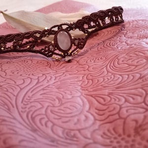 May include: A brown macrame headband featuring a central oval-shaped light-colored stone and small metallic gold beads woven throughout the corded design. The accessory has a symmetrical pattern with intricate knots and a pointed center.