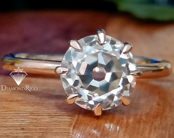 May include: A rose gold engagement ring with a round, clear, faceted gemstone set in a six-prong setting. The ring is on a wooden surface.