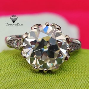 May include: A silver ring with a large, clear, round diamond in a prong setting. The diamond has many facets and sparkles brightly. The ring has smaller diamonds on the band.