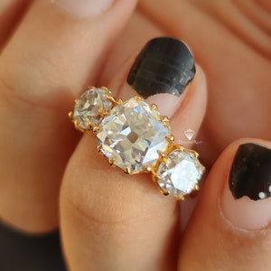 Puede incluir: Un anillo de oro con tres diamantes grandes, transparentes y de corte cuadrado. El anillo est&aacute; en un dedo con esmalte de u&ntilde;as negro.