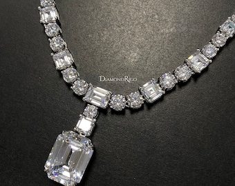 May include: A dazzling diamond necklace featuring a large emerald-cut center stone, complemented by a series of round and square-cut diamonds. The necklace is set in a silver metal and is displayed against a dark background. The brand name "DiamondRico" is visible.