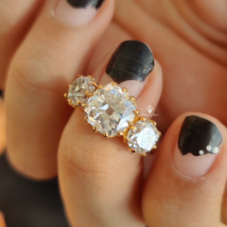 Puede incluir: Un anillo de oro con tres diamantes grandes, transparentes y de corte cuadrado. El anillo est&aacute; en un dedo con esmalte de u&ntilde;as negro.