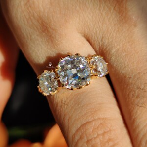 Puede incluir: Un anillo de oro con tres piedras preciosas grandes, claras y de corte cuadrado. El anillo est&aacute; en un dedo.