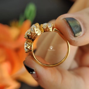 Puede incluir: Un anillo de oro con tres piedras preciosas grandes y transparentes engastadas en un dise&ntilde;o de corona. El anillo est&aacute; en un dedo y el nombre de la marca "DiamondRico" es visible dentro del anillo.