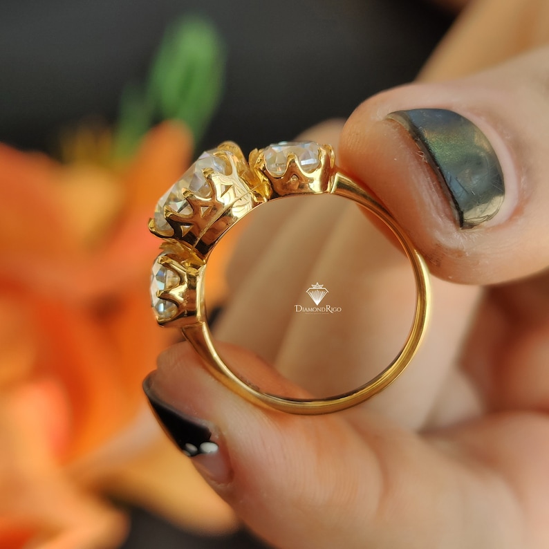 Puede incluir: Un anillo de oro con tres piedras preciosas grandes y transparentes engastadas en un dise&ntilde;o de corona. El anillo est&aacute; en un dedo y el nombre de la marca "DiamondRico" es visible dentro del anillo.