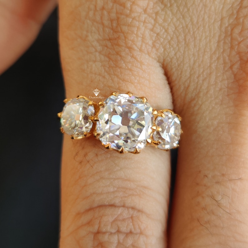 Puede incluir: Un anillo de oro con tres diamantes grandes, redondos y transparentes. Los diamantes est&aacute;n engastados en garras y est&aacute;n rodeados de diamantes m&aacute;s peque&ntilde;os.