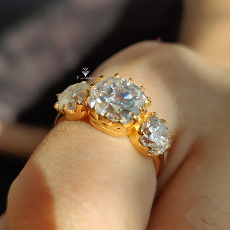Puede incluir: Un anillo de oro con tres piedras preciosas redondas y transparentes de gran tama&ntilde;o. El anillo est&aacute; en un dedo.