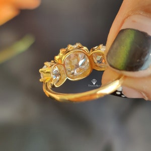 Puede incluir: Un anillo de oro con tres piedras preciosas redondas y transparentes engastadas en garras. El anillo est&aacute; en un dedo.