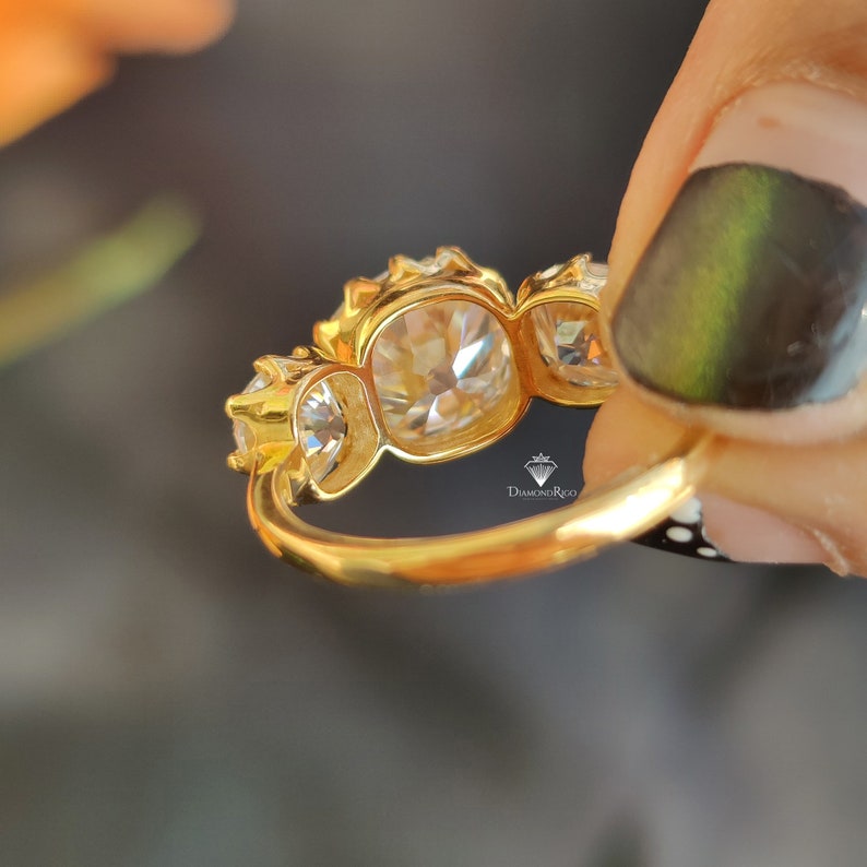 Puede incluir: Un anillo de oro con tres piedras preciosas redondas y transparentes engastadas en garras. El anillo est&aacute; en un dedo.