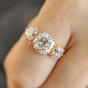 Puede incluir: Un anillo de oro con tres piedras preciosas grandes, redondas, transparentes y facetadas. El anillo est&aacute; en un dedo.