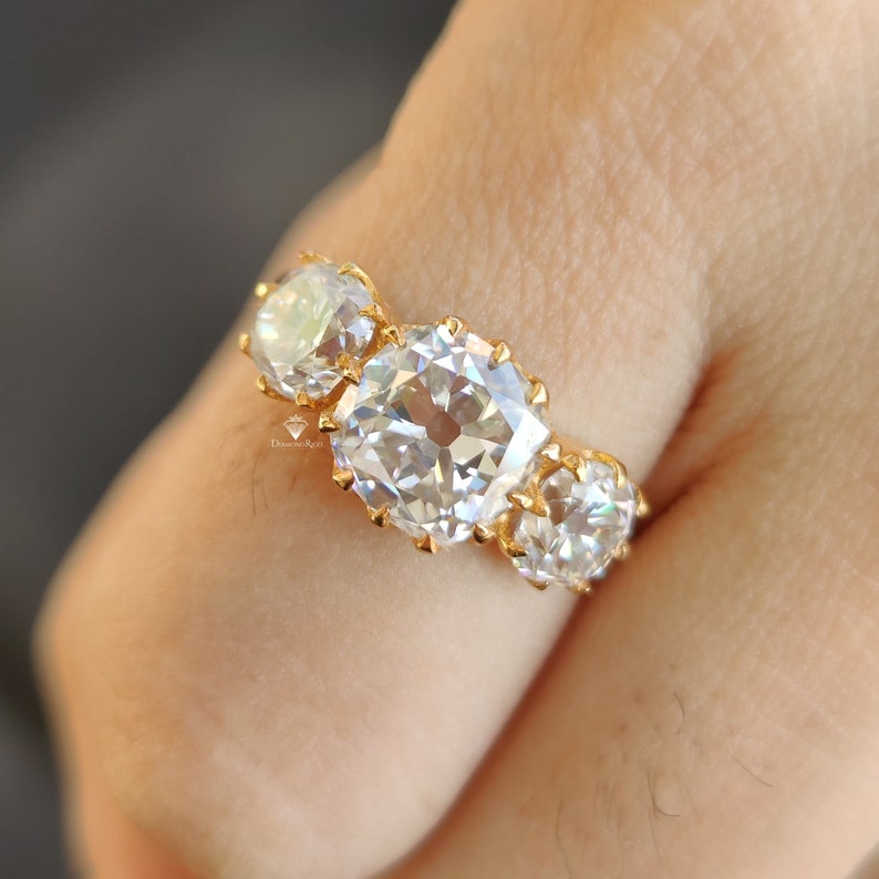 Puede incluir: Un anillo de oro con tres piedras preciosas grandes, redondas, transparentes y facetadas. El anillo est&aacute; en un dedo.
