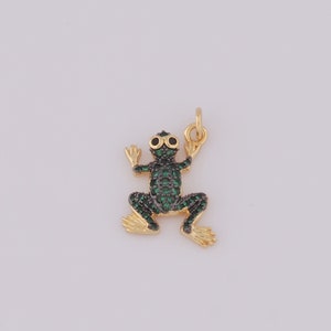 May include: A gold-colored frog charm with green and black accents. The frog's body is covered in small green stones, with black stones for the eyes. The charm has a loop at the top for attaching to a necklace or bracelet.