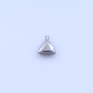 Puede incluir: Un pequeño colgante triangular plateado con un lazo en la parte superior para sujetarlo a una cadena o cordón. El colgante tiene una superficie lisa y pulida y una forma geométrica. El fondo es de color azul claro liso.