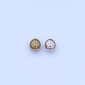 May include: Two small, turtle shell-shaped beads. One bead is gold-toned, the other is silver-toned. Each bead has a detailed, geometric pattern. The beads are displayed on a light blue background.