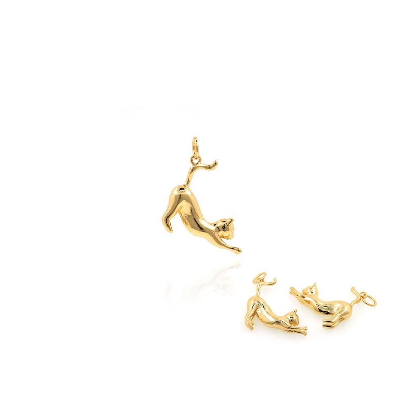 May include: Three gold-toned cat charms. The top charm is a pendant with a loop for a chain. The bottom two charms are smaller and are laying flat. All three charms are in the shape of a cat stretching.
