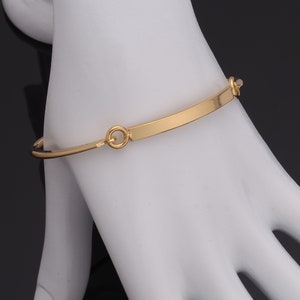Pode incluir: Uma pulseira de bracelete dourada com um design de barra retangular. A pulseira é feita de um metal brilhante e liso e apresenta um estilo simples e elegante. A pulseira é mostrada em uma superfície branca.