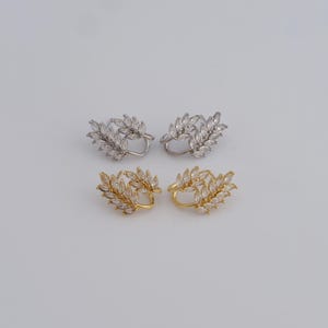 May include: Four ear cuff earrings. Two are silver-toned, and two are gold-toned. Each earring features a leaf design with clear, sparkling crystals. The earrings are arranged in pairs, showcasing their design and color variations.
