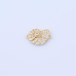 May include: A gold-colored jewelry clasp in the shape of a stylized flower. The clasp is encrusted with small, clear crystals, and has two small loops on either side for attachment. The flower design features a central crystal and radiating petals.
