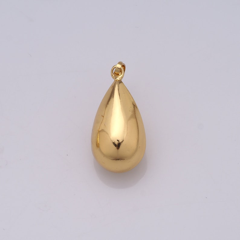 May include: A gold-colored teardrop pendant with a smooth, reflective surface. The pendant has a small loop at the top for a chain. The teardrop shape is rounded and symmetrical, reflecting light.