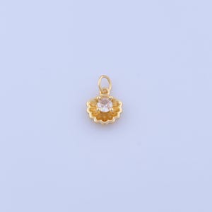 May include: A gold-toned metal charm shaped like a scalloped shell, featuring a small circular jump ring at the top and a single clear, round-cut gemstone set securely in the center of the textured, fan-like metal frame.