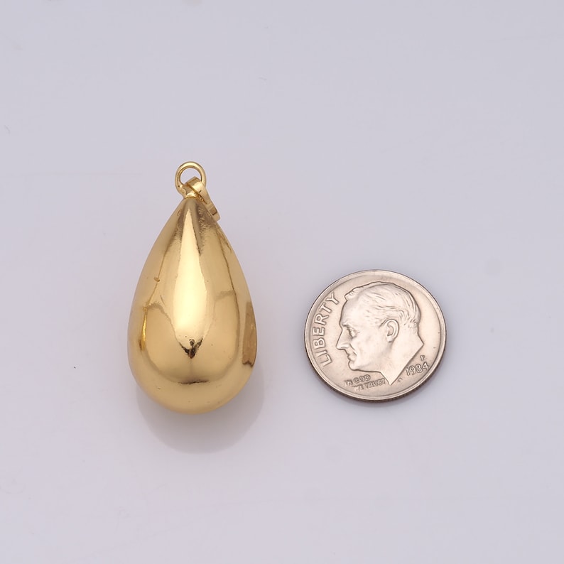 May include: A gold-colored teardrop pendant with a small loop at the top for a chain. The pendant is next to a U.S. dime for size comparison. The pendant has a smooth, reflective surface.