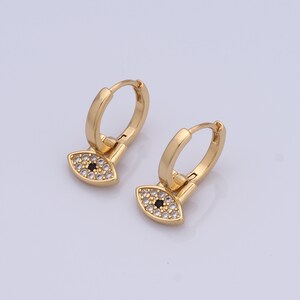 May include: Gold hoop earrings with a dangling evil eye charm. The charm is decorated with small white stones and a black centre.