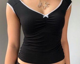 May include: Black cap-sleeve top with a V-neckline trimmed with white lace. A small white bow accents the gathered bust. Two gold necklaces with initial pendants complete the look. The top is form-fitting and appears to be made of a stretchy material.
