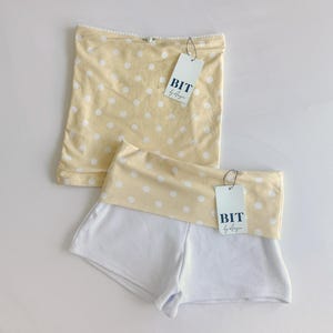 May include: Two-piece set of clothing. The top is a light yellow tube top with white polka dots. The bottom is white shorts with a light yellow polka dot waistband. Each piece has a tag that says "BIT by Angie".