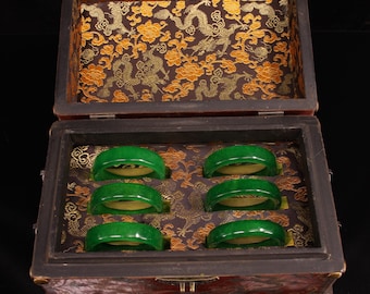 Puede incluir: Una caja de madera con tapa con bisagras, forrada con una tela dorada y verde con un diseño de dragón. La caja contiene seis pulseras de jade verde.