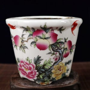 Könnte beinhalten: Ein weißer Keramiktopf mit einem Blumendesign mit rosa Pfingstrosen, gelben Blumen und Pfirsichzweigen. Der Topf hat einen gewellten Rand und einen leicht ausgestellten Boden.