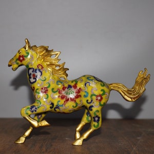 Puede incluir: Una figurita de caballo de cloisonné amarillo con intrincados patrones florales en tonos de azul, rojo, verde y blanco. El caballo está corriendo con la cola y la crin al viento.
