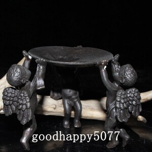 May include: A black metal candle holder with three cherubs holding up a round tray. The cherubs have wings and are in a standing position.