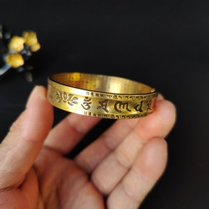 Può includere: Un braccialetto in metallo dorato con un'iscrizione in una lingua straniera. L'iscrizione include la parola "Om" e un motivo floreale.