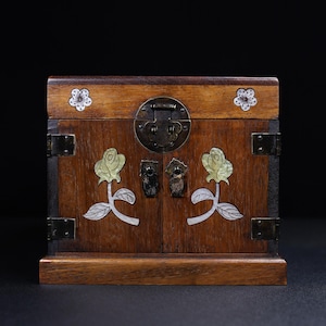 Peut inclure: Un coffre en bois avec un couvercle à charnière et deux portes. Le coffre est décoré de motifs floraux incrustés et de ferrures métalliques. Le bois a une teinte brune chaude, et le coffre semble être d'origine asiatique.