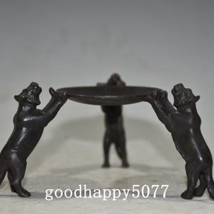 May include: A bronze sculpture of three stylized animals holding up a circular tray. The animals are in a seated position with their front paws raised to support the tray. The sculpture is a dark brown color and has a smooth finish.