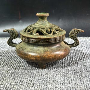 May include: A bronze-toned metal incense burner featuring a rounded body, three small feet, and two curved side handles. The removable lid has an openwork scroll design and a central knob, while the rim is decorated with a geometric Greek key pattern.