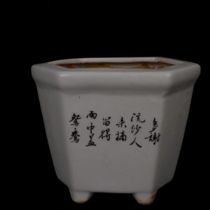 Pode incluir: Um vaso hexagonal de cerâmica branca com uma inscrição preta em caracteres chineses. O vaso tem três pequenos pés.