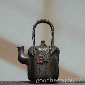 May include: A dark metallic teapot with a textured, rope-style handle. The body features sculpted green leaves and a single reddish-brown flower. The text "goodhappy5077" is visible at the bottom. The spout is short and curved, and the lid has a small knob.