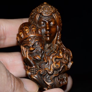 Puede incluir: Una escultura de madera tallada a mano de una mujer con una máscara. La mujer lleva una corona y tiene los ojos cerrados. La máscara es un demonio con una expresión feroz. La escultura está intrincadamente detallada y tiene un rico color marrón.