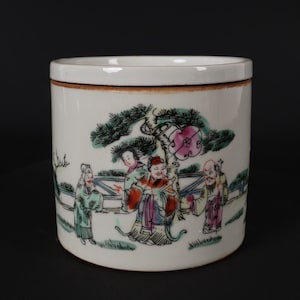 Könnte beinhalten: Ein weißer zylindrischer Behälter aus Keramik mit Deckel, der eine handgemalte Szene mit vier Figuren in einer Landschaft mit einer Kiefer zeigt. Die Figuren sind in farbenfrohe Roben gekleidet und in einem verspielten, fantasievollen Stil dargestellt.