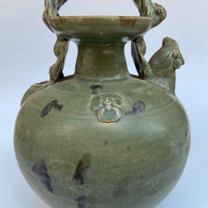 May include: A celadon green ceramic vessel with a rounded body, a wide neck, and a curved handle arching over the top. The piece features a sculpted bird figure on the shoulder and a small floral relief detail on the front, with dark brown spots.
