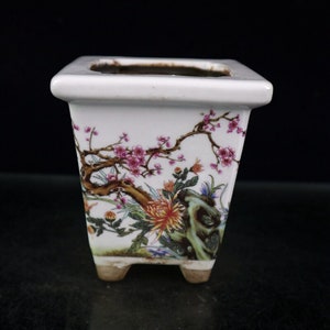 Könnte beinhalten: Ein weißer quadratischer Keramiktopf mit Blumenmuster. Der Topf hat einen quadratischen Boden und vier kleine Füße. Das Design zeigt rosa und weiße Blumen, grüne Blätter und braune Zweige.