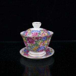 Pode incluir: Um conjunto de chávena e pires de porcelana branca com um padrão floral em tons de rosa, azul, amarelo e verde. A chávena tem uma tampa e uma borda dourada.