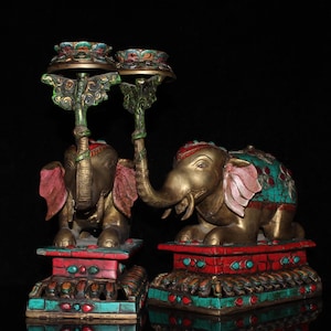 May include: Two antique brass elephant candle holders. Each elephant has a trunk raised and is adorned with red, turquoise, and gold accents. The candle holders are on a red and turquoise base, with a black background.