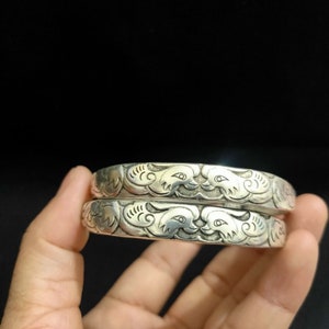 Puede incluir: Dos brazaletes de plata con intrincados diseños grabados de pájaros y nubes estilizados.