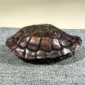 May include: A bronze-coloured turtle shell sculpture. The shell has a segmented pattern, with a polished, reflective surface. The sculpture is likely a decorative item or collectible, possibly for display.