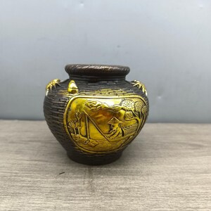 Puede incluir: Un jarrón decorativo de color bronce con un panel dorado que presenta una escena grabada de una persona pescando desde un barco. El jarrón tiene una textura tejida y pequeños detalles dorados.