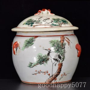 May include: A white ceramic jar with a matching lid, featuring hand-painted illustrations of cranes and pine trees in green, black, and red. The jar has two red side handles and a red-trimmed rim, with a rounded body and a domed lid top.